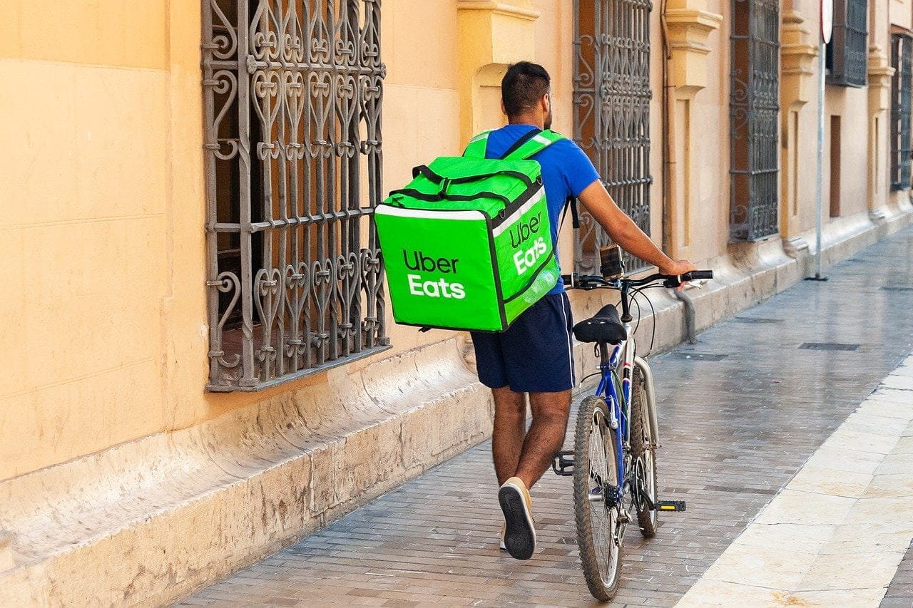 Com caixa de entrega nas costas, com logomarca da Uber Eats, entregador caminha sozinho por calçada enquanto empurra a bicicleta.