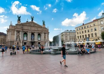 Fachada do teatro ópera e balé diante da praça onde é possível ver uma fonte lançando água para o alto e pessoas caminhando pela praça em Lviv, na Ucrânia.