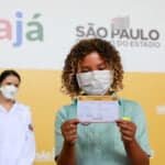 Criança negra, usando máscara de proteção facial, segura cartão de vacinação diante de um painel que traz a imagem de uma enfermeira, a logomarca do Estado e a palavra "vacinajá".