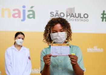 Criança negra, usando máscara de proteção facial, segura cartão de vacinação diante de um painel que traz a imagem de uma enfermeira, a logomarca do Estado e a palavra "vacinajá".