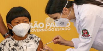 Davi, garoto de origem indígena, usando máscara, recebe a vacina, aplicada por uma enfermeira de jaleco branco.