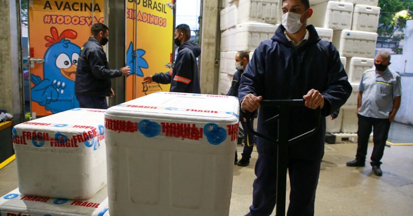 Funcionário do Instituto Butantan transporta em um carrinho de mão caixas de isopor com doses da Coronavac. Ao fundo, um caminhão com o baú envelopado com o desenho de uma galinha em cor azul e fundo amarelo.