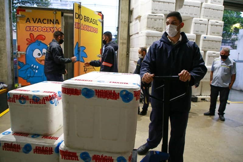Funcionário do Instituto Butantan transporta em um carrinho de mão caixas de isopor com doses da Coronavac. Ao fundo, um caminhão com o baú envelopado com o desenho de uma galinha em cor azul e fundo amarelo.