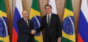 Vladimir Putine Jair Bolsonaro se cumprimentam dando a mão diante de bandeiras dos dois países.