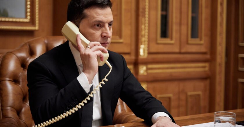 Volodimyr Zelensky, presidente da Ucrânia, fala ao telefone sentado à mesa. A mão esquerda está estendida sobre a mesa, onde há folhas de papeis.