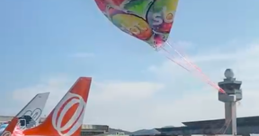 Balão colorido com imagens de personagens do SBT se aproxima do chão enquanto outra parte já está sobre a calda de um avião estacionado.
