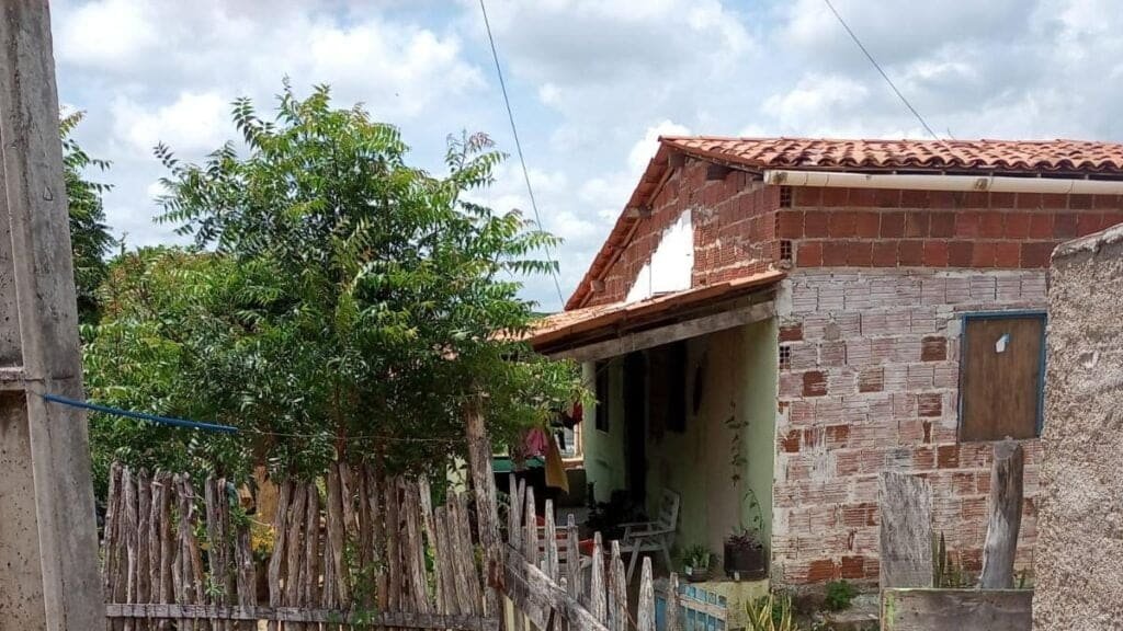 Foto mostra parte de fora de uma casa em Canindé, ceará, com paredes sem reboco. É possível ver também uma árvore em frente a casa e uma cerca de madeira. 