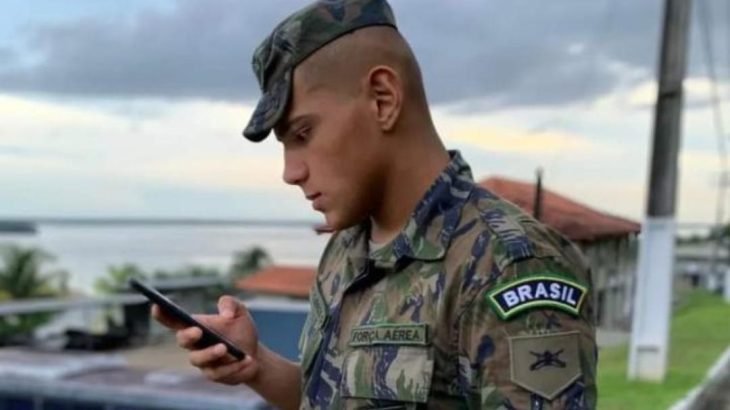 Gustavo Henrique Souza Gomes, usando farda, e boné na cabeça, segura o celular com a mão direita próximo ao rosto enquanto olha para o aparelho. 