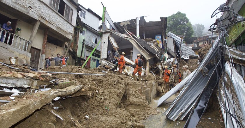 Foto mostra rua tomada pela terra e bombeiros sobre a lama trabalhando. É possível ver ferros retorcidos e casas destruídas em Petrópolis.
