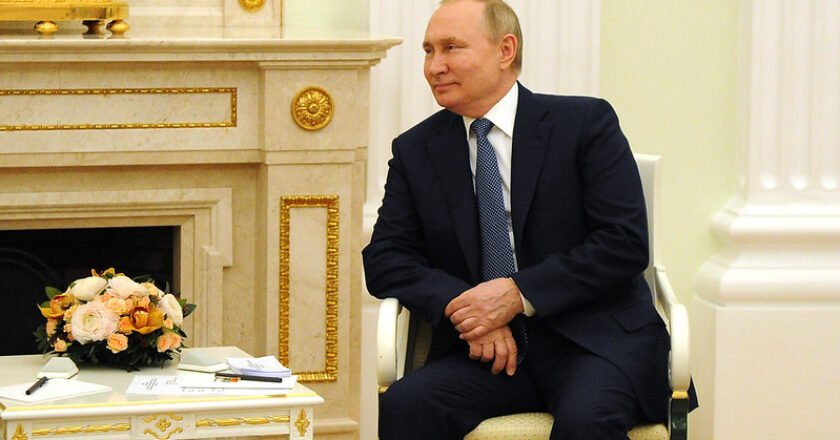 Vladimir Putin, presidente da Rússia, sentado, veste terno e gravata, com paletó aberto e as duas mãos cruzadas sobre as pernas. É possível ver uma mesa de centro um flores em cima e papeis, além de uma lareira ao fundo.