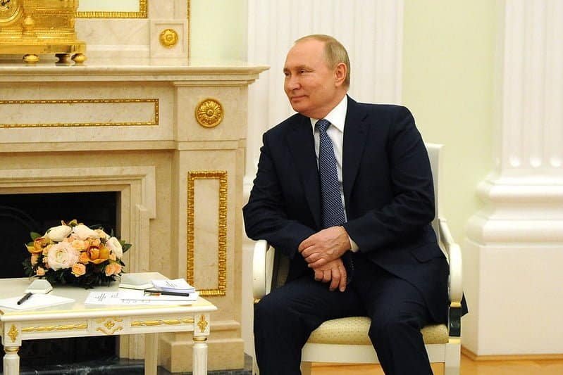 Vladimir Putin, presidente da Rússia, sentado, veste terno e gravata, com paletó aberto e as duas mãos cruzadas sobre as pernas. É possível ver uma mesa de centro um flores em cima e papeis, além de uma lareira ao fundo. 