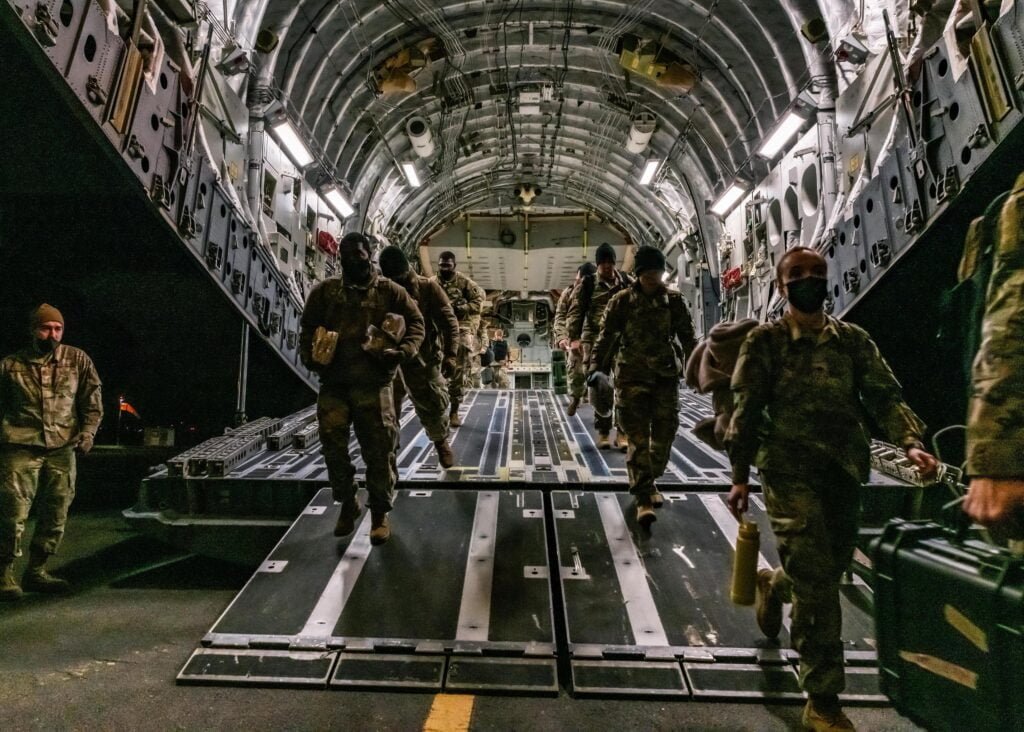 Soldados descem de uma aeronave miliar. Todos usam farda. Desembarque acontece à noite.