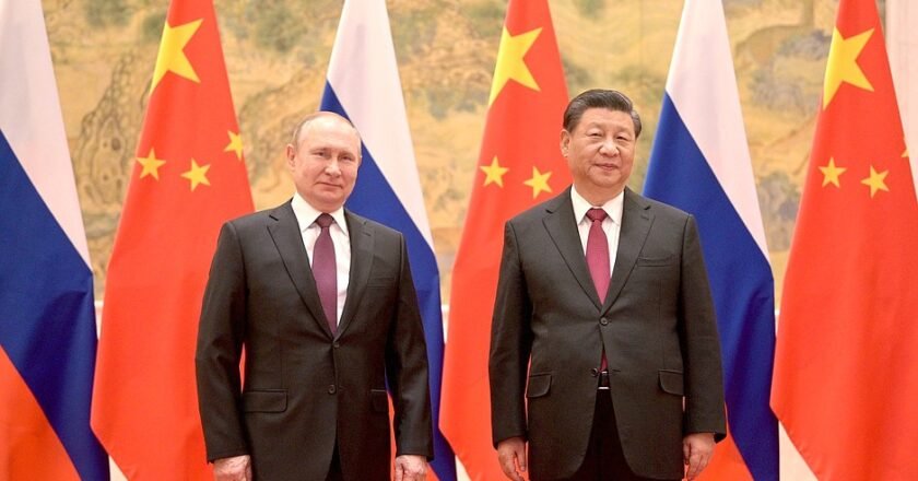 Vladimir Putin, homem branco e cabelos claros, ao lado de Xi Jinpin, homem de pele clara, olhos puxados. Os dois usam terno e gravata e posam para foto tendo bandeiras dos dois países ao fundo.
