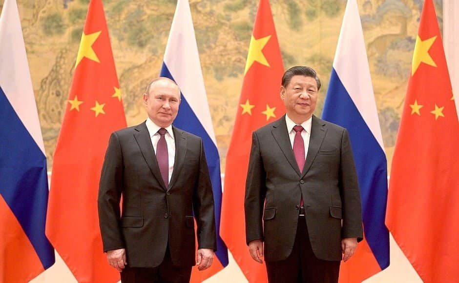 Vladimir Putin, homem branco e cabelos claros, ao lado de Xi Jinpin, homem de pele clara, olhos puxados. Os dois usam terno e gravata e posam para foto tendo bandeiras dos dois países ao fundo. 