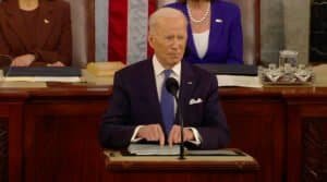 Presidente Joe Biden, de terno e gravata, diante de um microfone, aponta os dedos indicadores para baixo, tocando o púlpito de onde discursa