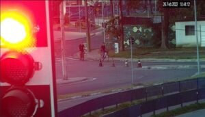Foto mostram em primeiro plano um semáforo de trânsito e, ao fundo, dois homens em bicicletas. É possível ver também imóveis da região e cones de sinalização perto da calçada. Imagem mostra minutos antes da abordagem.