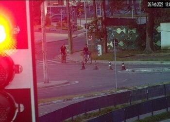 Foto mostram em primeiro plano um semáforo de trânsito e, ao fundo, dois homens em bicicletas. É possível ver também imóveis da região e cones de sinalização perto da calçada. Imagem mostra minutos antes da abordagem.