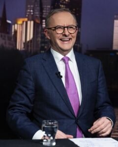 Anthony Albanese, homem de pele clara, óculos de grau arredondado, sorri enquanto concede entrevista em um estúdio, vestindo terno e gravata. 