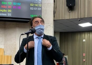 Adilson Amadeu, de terno e gravata, usando máscara, discursa ao microfone da Câmara Municipal enquanto aponta os dois dedos indicadores para si mesmo.