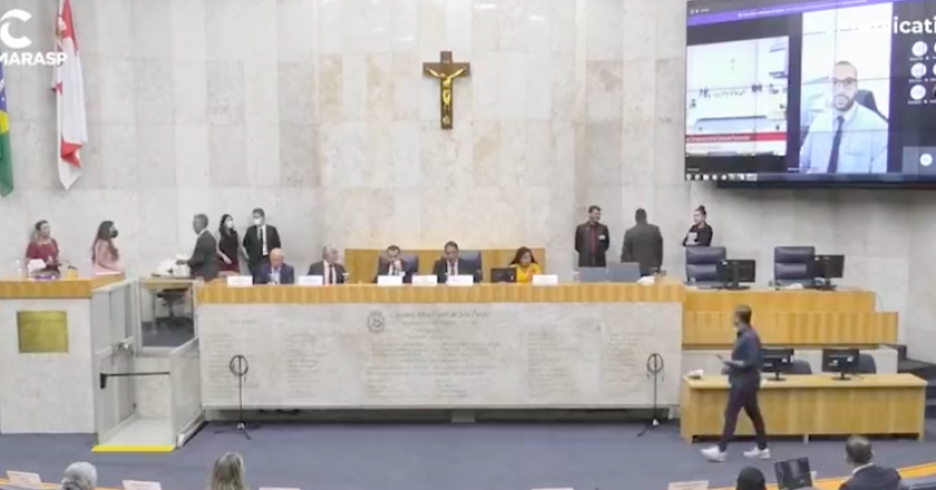 Plenário da Câmara municipal de São Paulo. Parlamentares sentados à mesa diretora enquanto assessores caminham pelo local. No telão a imagem de um vereador.