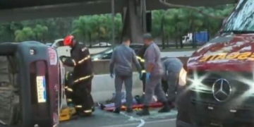 Carro tombado no asfalto, bombeiros socorrendo vítima e parte da frente da ambulância de resgate no canto direito da imagem.