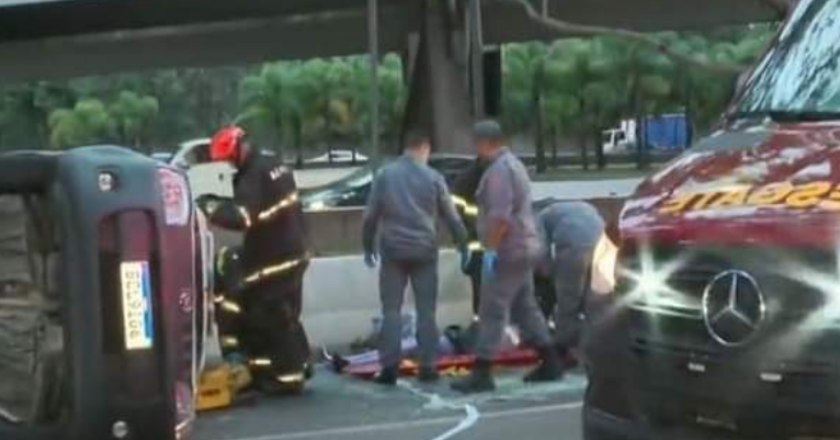 Carro tombado no asfalto, bombeiros socorrendo vítima e parte da frente da ambulância de resgate no canto direito da imagem.