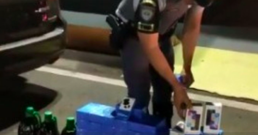 Policial Militar apresenta celulares encontrados dentro de carro. Caixas são empilhadas no chão.