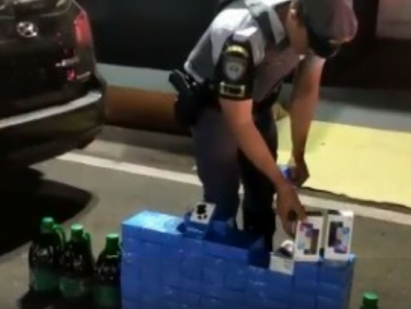 Policial Militar apresenta celulares encontrados dentro de carro. Caixas são empilhadas no chão.