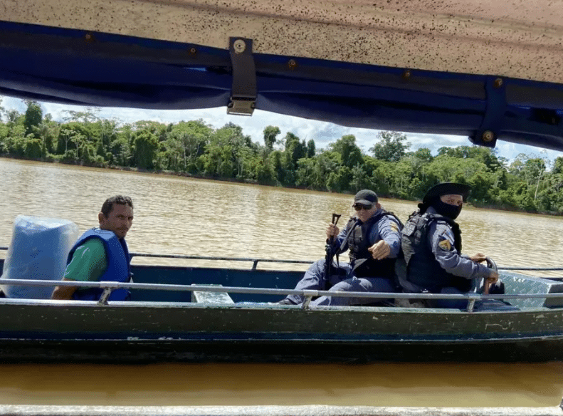 Amarildo da Costa de Oliveira, conhecido como pelado, em barco com dois policiais. Ele aparece com as mãos para trás.