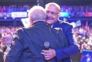 Geraldo Alckmin abraça sorrindo Lula, que aparece de costas. Ao fundo, uma arquibancada cheia de gente.