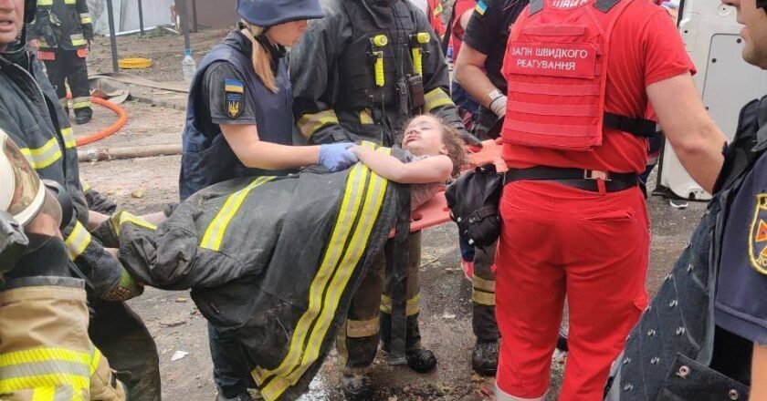 Criança deitada em maca e coberta com uniforme de bombeiro é carregada por socorristas após ataque.