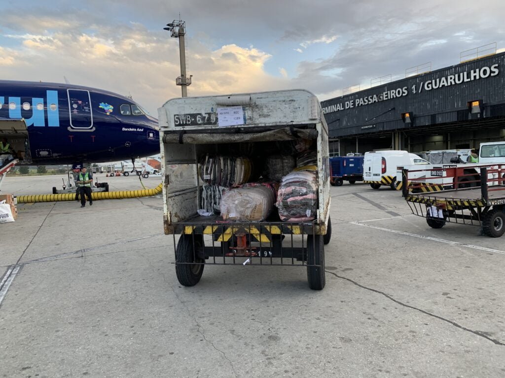 Carrinho de transporte de malas em aeroportos cheio de doações, indo em direção à aeronave no aeroporto de Guarulhos. No canto esquerdo aparece o bico do avião. 