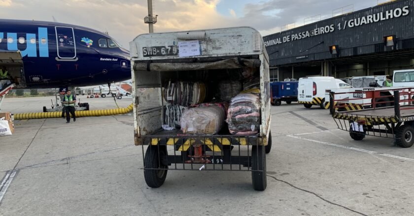 Carrinho de transporte de malas em aeroportos cheio de doações, indo em direção à aeronave no aeroporto de Guarulhos. No canto esquerdo aparece o bico do avião.