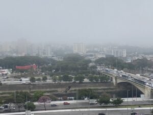Céu coberto por nuvens. Nevoeiro sobre os prédios da zona norte de São Paulo. Na parte de baixo da foto, a Marginal TietÊ com vários carros passando.