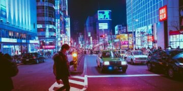 Homem atravessa a rua sobre a faixa de pedestres durante a noite enquanto carros aguardam. No letreiro, no canto direito da imagem, é possível ver o nome de uma loja escrito no idioma do Japão.