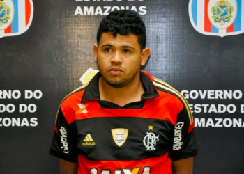 Luciano da Silva Barbosa, o "L7", diante do painel da secretaria de segurança pública do amazonas. Ele está com as mãos para trás e veste camisa de time de futebol. Aparece olhando para frente.