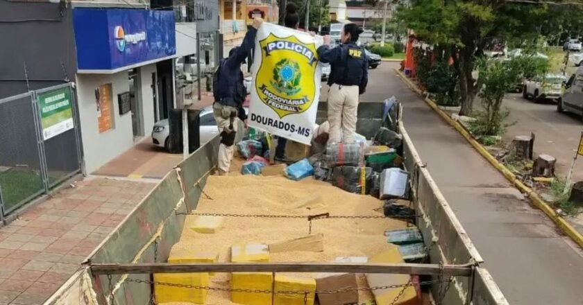 Policais sobre carroceria de caminhão onde droga foi encontrada. Eles seguram um baner com o símbolo da PRF e o nome da cidade de Dourados. A droga aparece em meio a grãos.