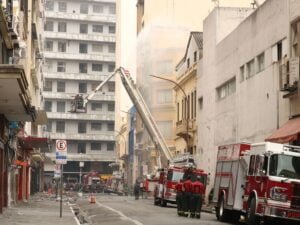 Caminhão dos bombeiros usa escada hidráulica para lançar água sobre o prédio atingido pelo fogo. Rua isolada tem apenas viaturas dos bombeiros e militares.