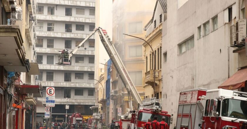 Caminhão dos bombeiros usa escada hidráulica para lançar água sobre o prédio atingido pelo fogo. Rua isolada tem apenas viaturas dos bombeiros e militares.