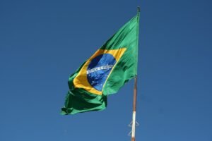 Bandeira do Brasil hasteada, na qual é possível ler Ordem e Progresso. Ao fundo, um céu azul