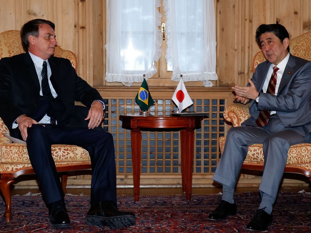 Jair Bolsonaro sentado ao lado de Shinzo Abe, durante encontro na Suíça. No centro, entre os dois, uma mesa tem bandeiras pequenas dos dois países.