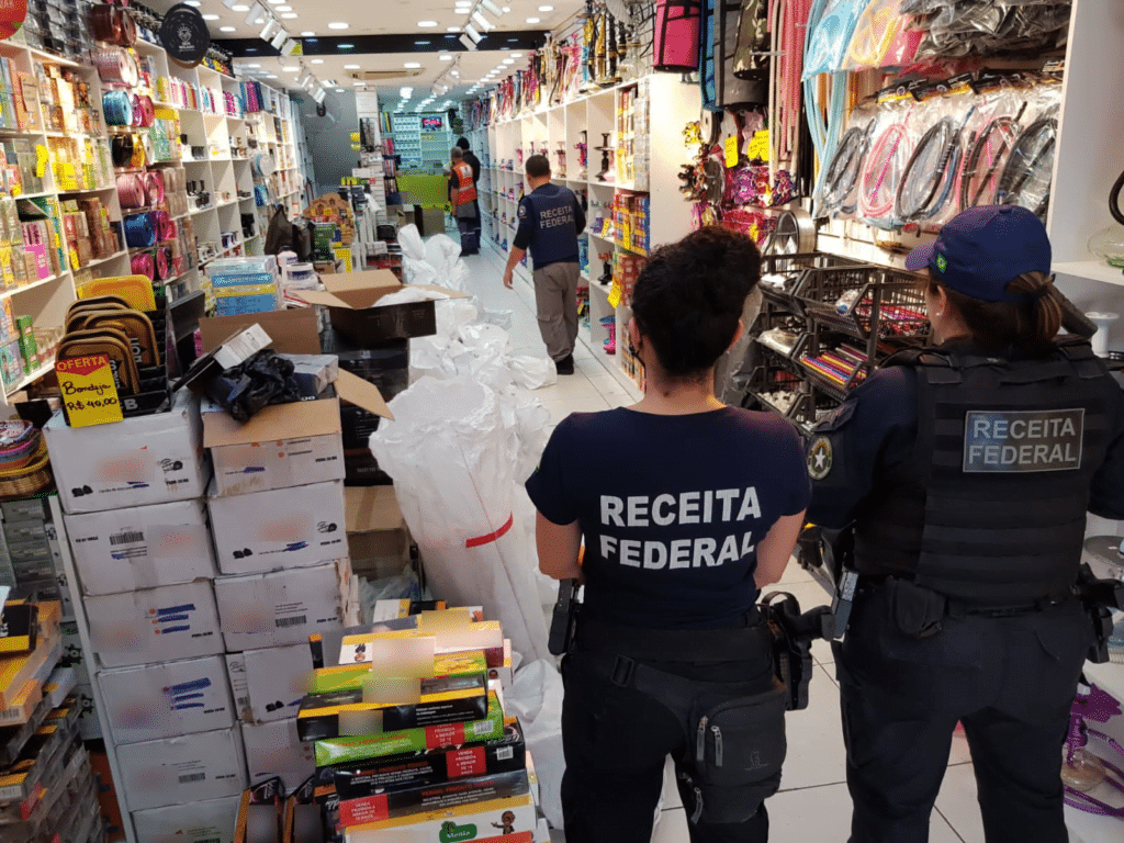 Agentes da Receita Federal, com o nome do órgão estampado na parte de trás da camiseta, observam loja cheia de mercadorias. 