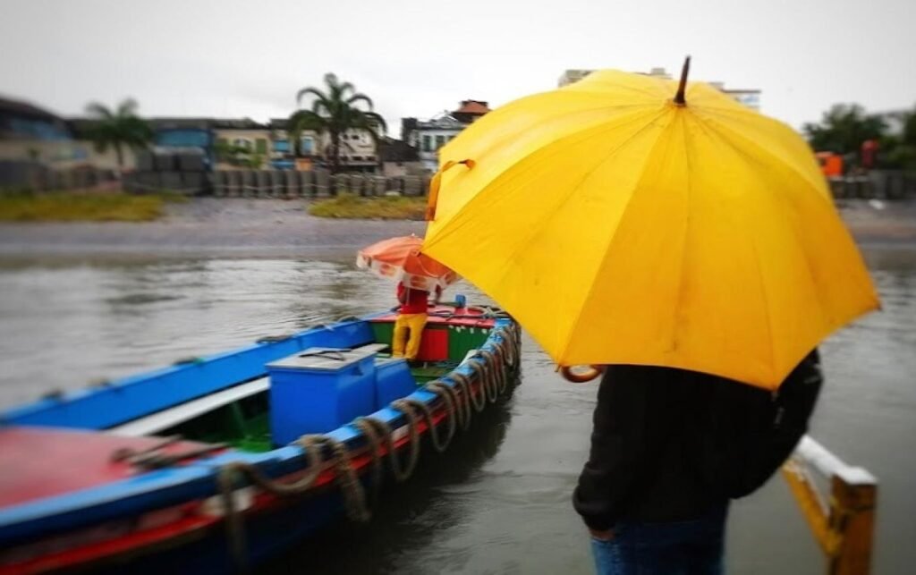 Pessoa usa guarda-chuvaao se aproximar de um barco, onde outra pessoa está dentro e também usando proteção para a chuva. Ao fundo, parte do mar sem ondas.
