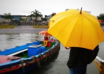 Pessoa usa guarda-chuvaao se aproximar de um barco, onde outra pessoa está dentro e também usando proteção para a chuva. Ao fundo, parte do mar sem ondas.