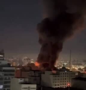 Fumaça e claridade provocada pelas chamas aparecem no meio de prédios em uma foto panorâmica.