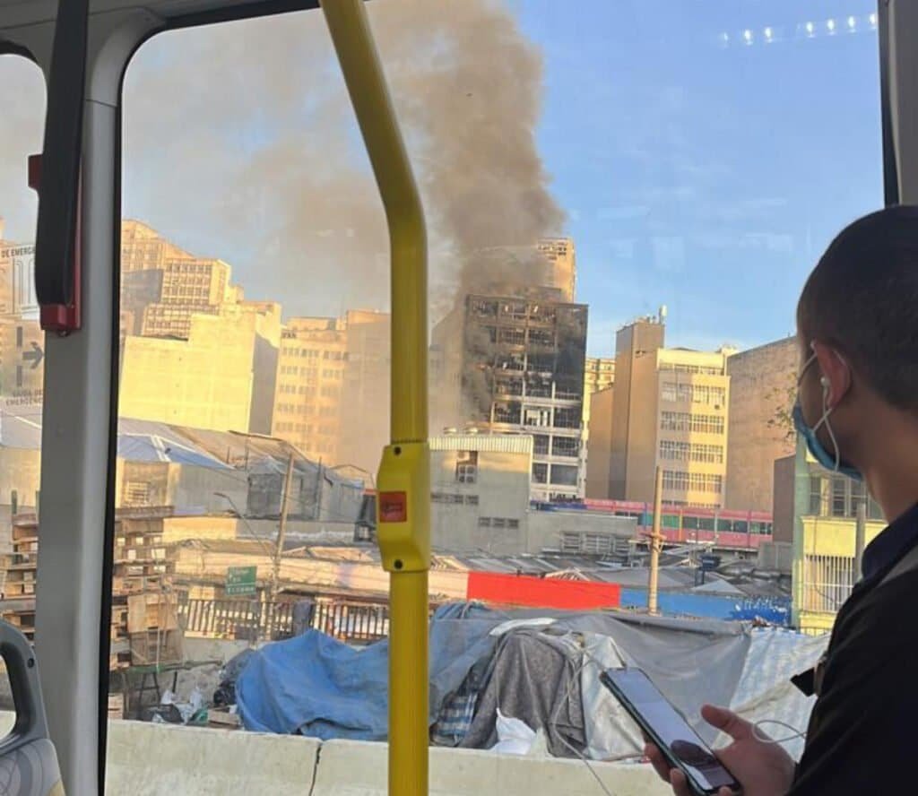 Dentro de ônibus, homem observa prédio destruído pelas chamas na região da 25 de março. É possível ver fumaça saindo do edifício.