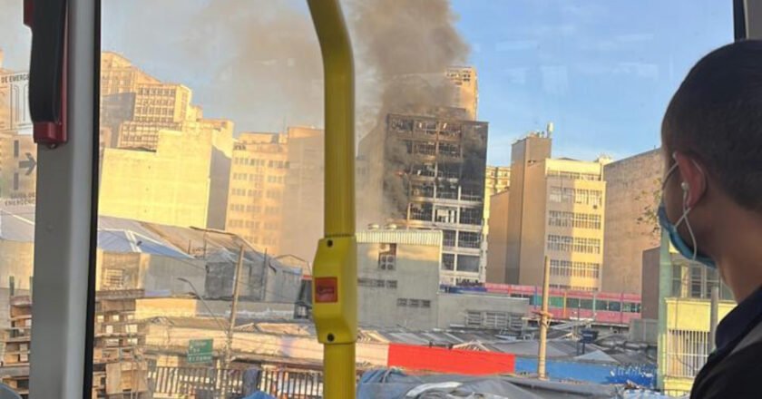 Dentro de ônibus, homem observa prédio destruído pelas chamas na região da 25 de março. É possível ver fumaça saindo do edifício.