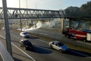 Bombeiros apagam fogo no meio da rodovia enquanto carros passam por outra pista ao lado.