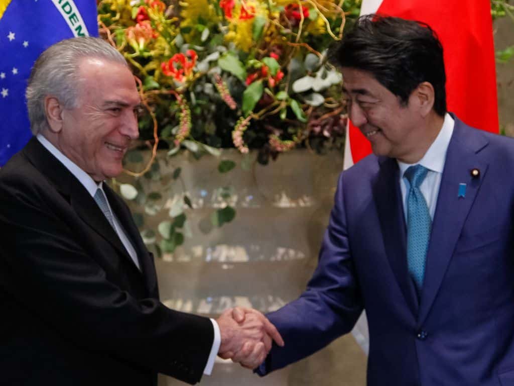Michel Temer, ex-presidente da República, cumprimenta Shinzo Abe durante visita ao Japão. Os dois sorriem enquanto dão as mãos. Ao fundo, bandeiras do Brasil e do Japão.