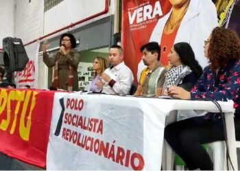 Vera Lúcia discursa ao microfone ao lado de uma mesa formada por homens e mulheres. Faixas com o nome da candidata aparecem no local.